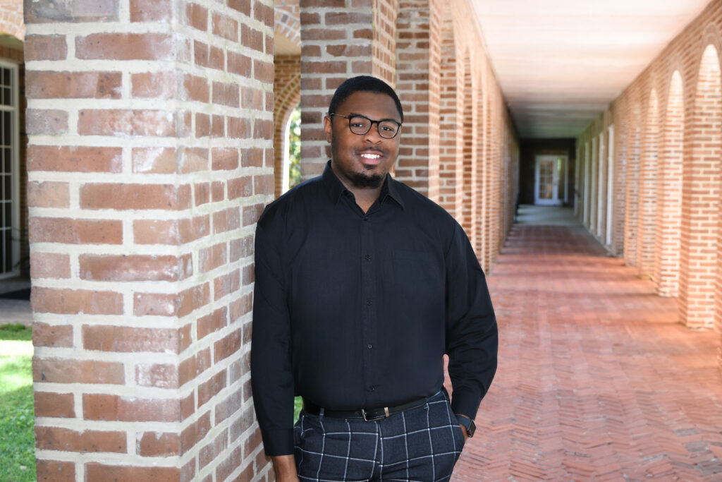 Student in glasses and black shirt and black plaid pants leans against a brick wall