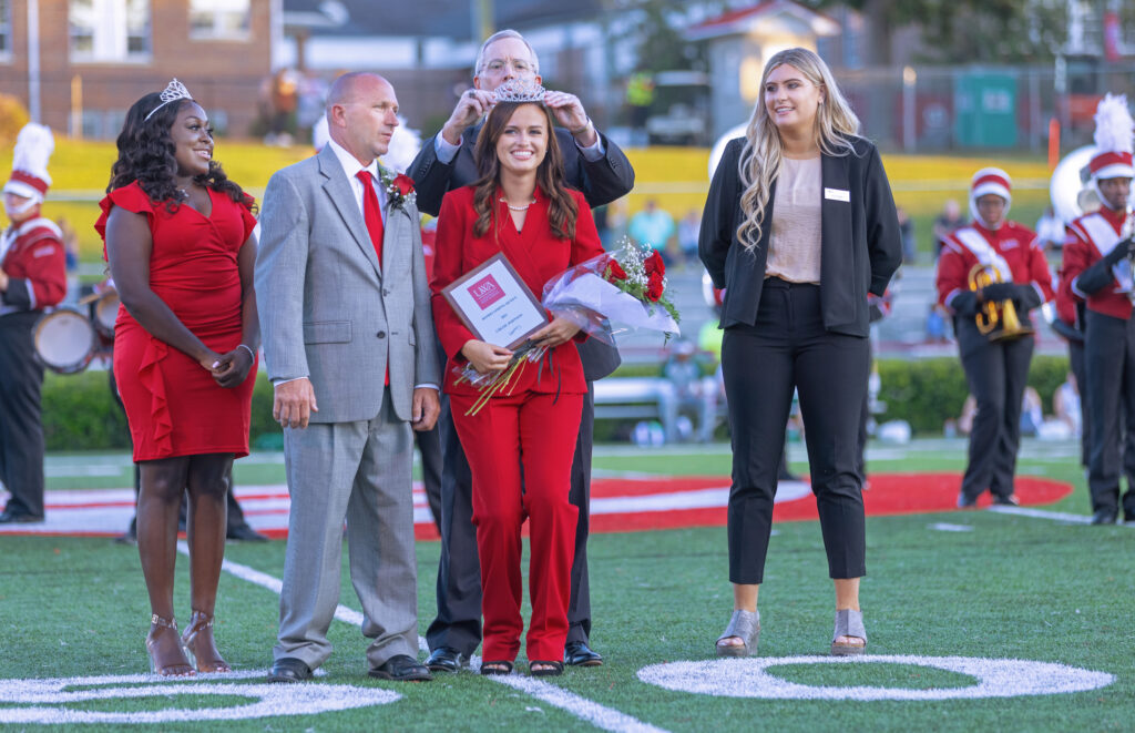 Brunette white female is crowned on the football field homecoming queen by UWA President Ken Tucker while her father and two others watch


